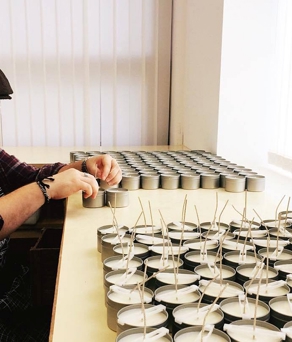 A bearded man, wearing a flat cap and an apron, sits at a desk adding wicks to candles.