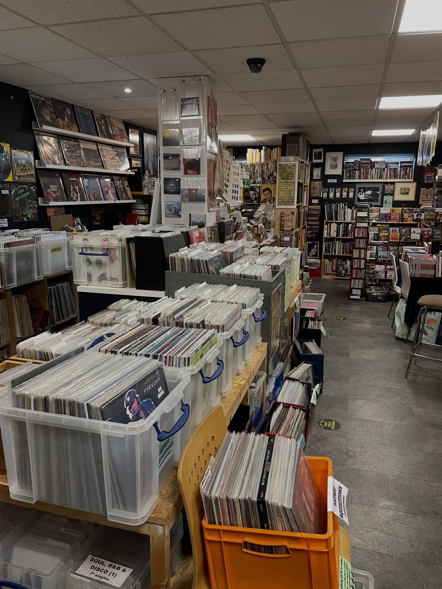 An eclectic shop interior combining vinyl records and books. Plastic crates filled with vinyl records dominate the foreground, arranged in rows on wooden tables. The background is lined with shelves of books and framed posters, creating a cozy, packed atmosphere. The floor is gray tile, and the ceiling has white panels with bright lighting.