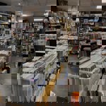 An eclectic shop interior combining vinyl records and books. Plastic crates filled with vinyl records dominate the foreground, arranged in rows on wooden tables. The background is lined with shelves of books and framed posters, creating a cozy, packed atmosphere. The floor is gray tile, and the ceiling has white panels with bright lighting.