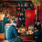 Three men are sat drinking in Hop Hideout, with a male bartender putting a drink down on their table. A female bartender is in the background at the bar on the left, with a shelving unit full of their stock to the right by the entrance.