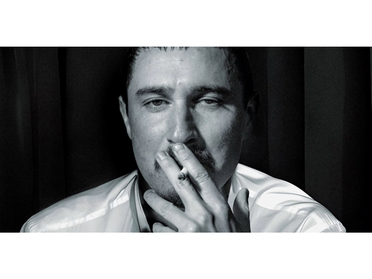 A person wearing a light-colored, long-sleeved shirt is seated against a dark background with vertical panels. One hand is raised near the chin, partially covering the mouth, creating a contemplative pose. The lighting emphasises the folds and sheen of the shirt, contrasting with the shadowed backdrop.