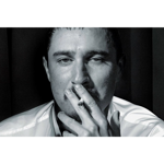 A person wearing a light-colored, long-sleeved shirt is seated against a dark background with vertical panels. One hand is raised near the chin, partially covering the mouth, creating a contemplative pose. The lighting emphasises the folds and sheen of the shirt, contrasting with the shadowed backdrop.