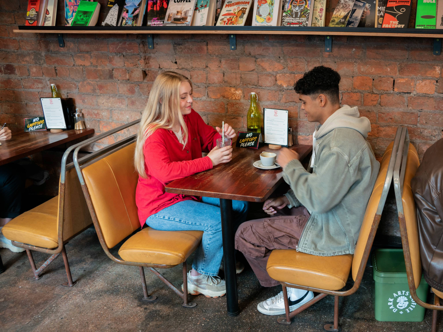 Two people are seated at a wooden table in a cosy café with exposed brick walls and tan leather booth seating. They have drinks in front of them, including a cup on a saucer and a glass with a straw. A green glass bottle and a small sign reading “NO LAPTOPS PLEASE” are on the table. Behind them, a shelf displays various colourful magazines and books. The café has a relaxed, informal atmosphere.