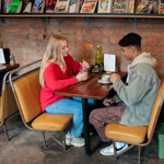 Two people are seated at a wooden table in a cosy café with exposed brick walls and tan leather booth seating. They have drinks in front of them, including a cup on a saucer and a glass with a straw. A green glass bottle and a small sign reading “NO LAPTOPS PLEASE” are on the table. Behind them, a shelf displays various colourful magazines and books. The café has a relaxed, informal atmosphere.