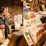 A split image; one side shows people browsing a stall, the other side shows a stall selling prints and cards.