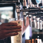 A close-up of a hand holding a plastic cup under a beer tap as a light, bubbly drink is poured.