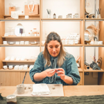 A person seated at a wooden table shaping a small handmade object, with tools and materials laid out in front, and shelves of ceramics and homewares displayed behind.