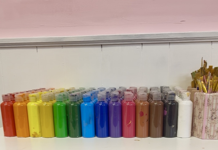 A row of small plastic paint bottles arranged by colour in a rainbow order on a white shelf, with brushes standing in a container at the far end against a pale pink and white wall.