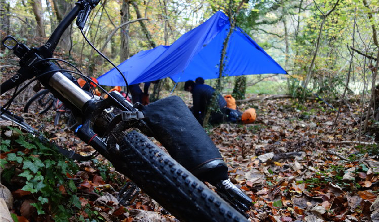 A shelter set up in a wooded area. A bike lies on the ground nearby. 