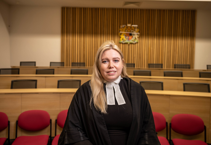 A person wearing a black robe and white neck bands is standing in a courtroom setting. Behind them are rows of red chairs and wooden benches, with a wooden panel wall featuring a coat of arms at the centre. The lighting is warm, and the room has a formal, official atmosphere.