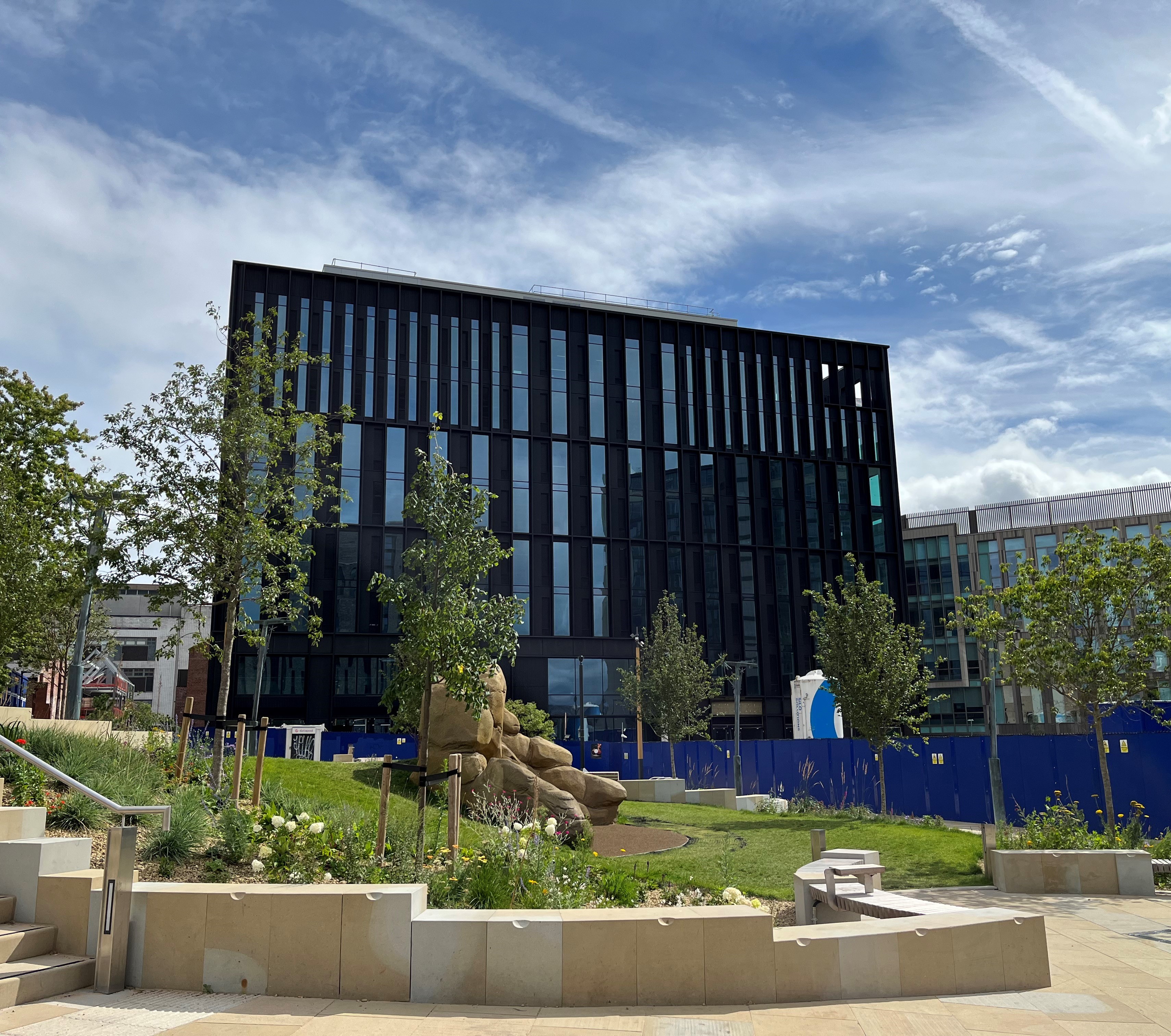 A modern black glass building with vertical panels stands behind a landscaped area featuring young trees, green grass, and large decorative rocks. Blue construction fencing surrounds part of the site, and the sky above is bright with scattered clouds and contrails.