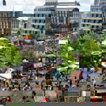 A wide aerial view of a food festival with colourful stalls, trees, and surrounding city buildings.