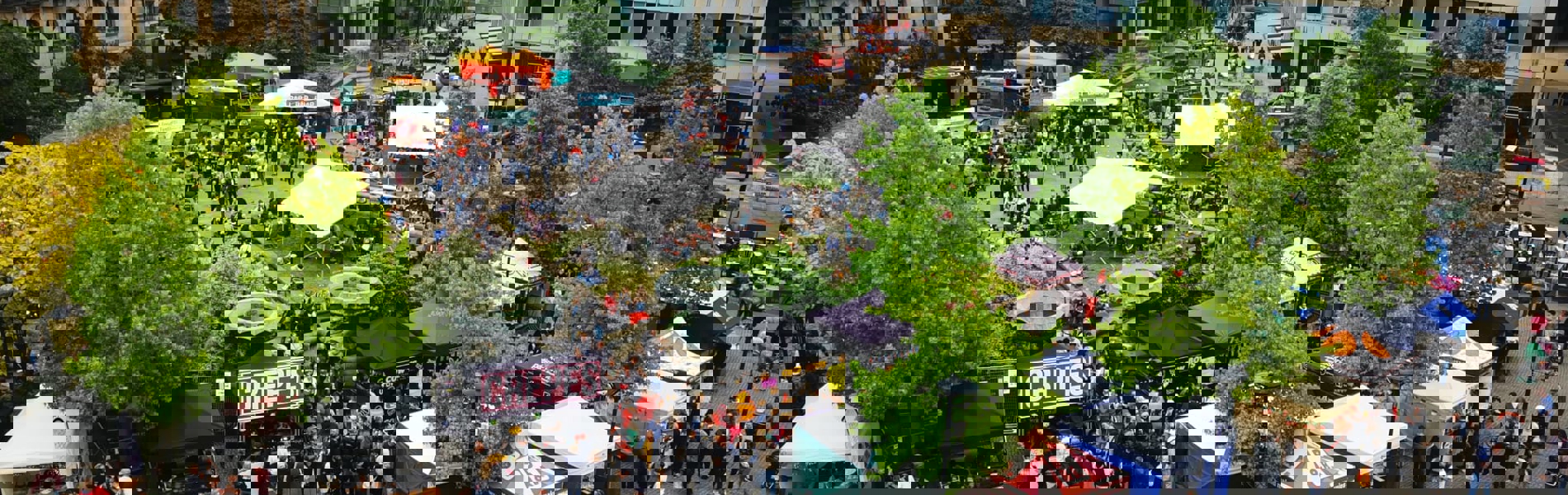 A wide aerial view of a food festival with colourful stalls, trees, and surrounding city buildings.