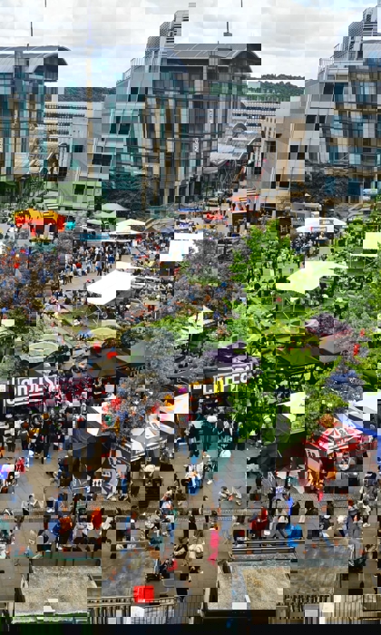 A wide aerial view of a food festival with colourful stalls, trees, and surrounding city buildings.