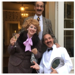 Three people are standing in front of an open doorway with glass panels. The person on the left is wearing a brown textured suit with a large black fur collar and a bright pink bow tie, holding a champagne flute in one hand and a handbag in the other. The person in the middle is dressed in a grey checked blazer with a white shirt and striped tie. The person on the right is wearing a white tuxedo jacket with a black bow tie and is holding a silver serving tray. 