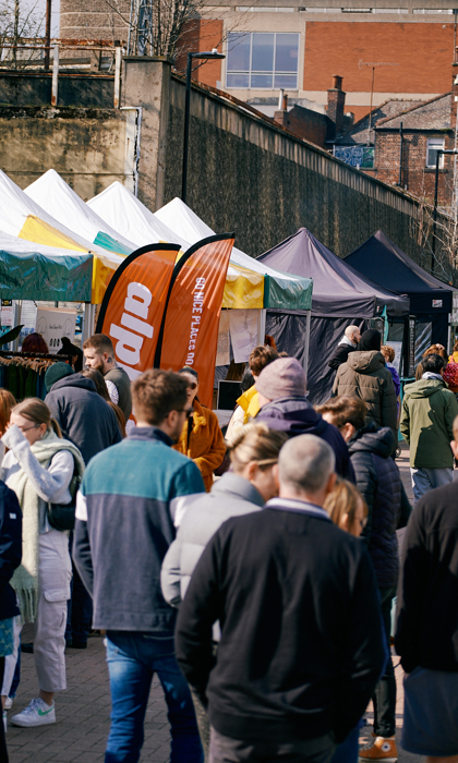 An open air market in a city centre. There are lots of people walking up and down and browsing the stalls.