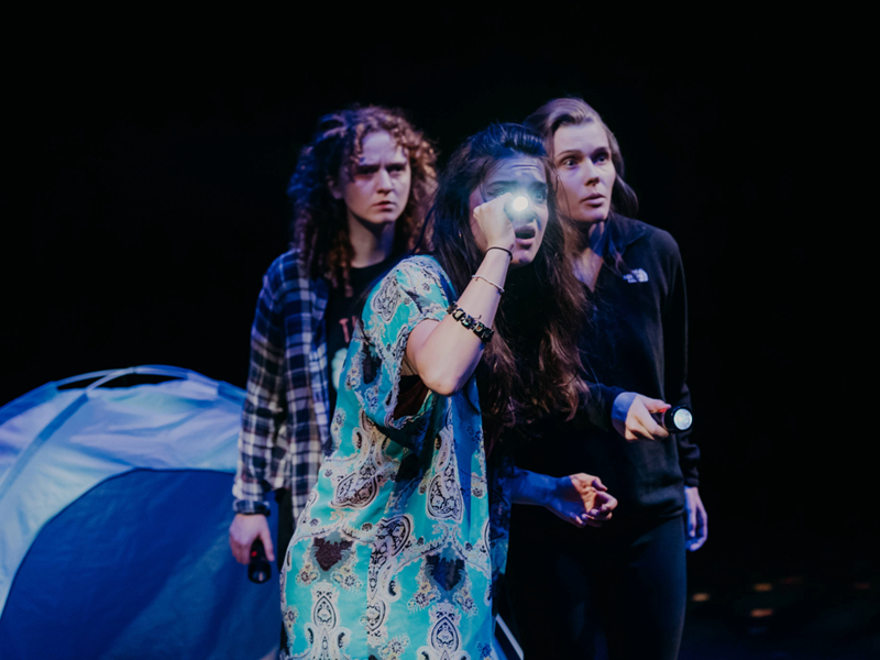 Three women, onstage as part of a play, are looking worried. Two of them are holding torches. Behind them is a blue tent.
