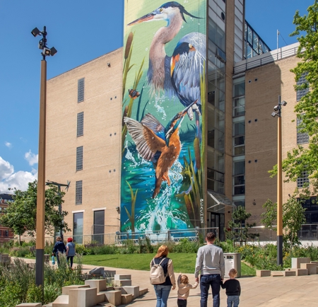 A large mural of two herons and a kingfisher in a vibrant wetland scene painted on the side of a tall modern building. In the foreground, a landscaped urban park with trees, benches, and pathways is visible, where several people, including a family with two children, are walking and enjoying the space under a bright blue sky with scattered clouds.