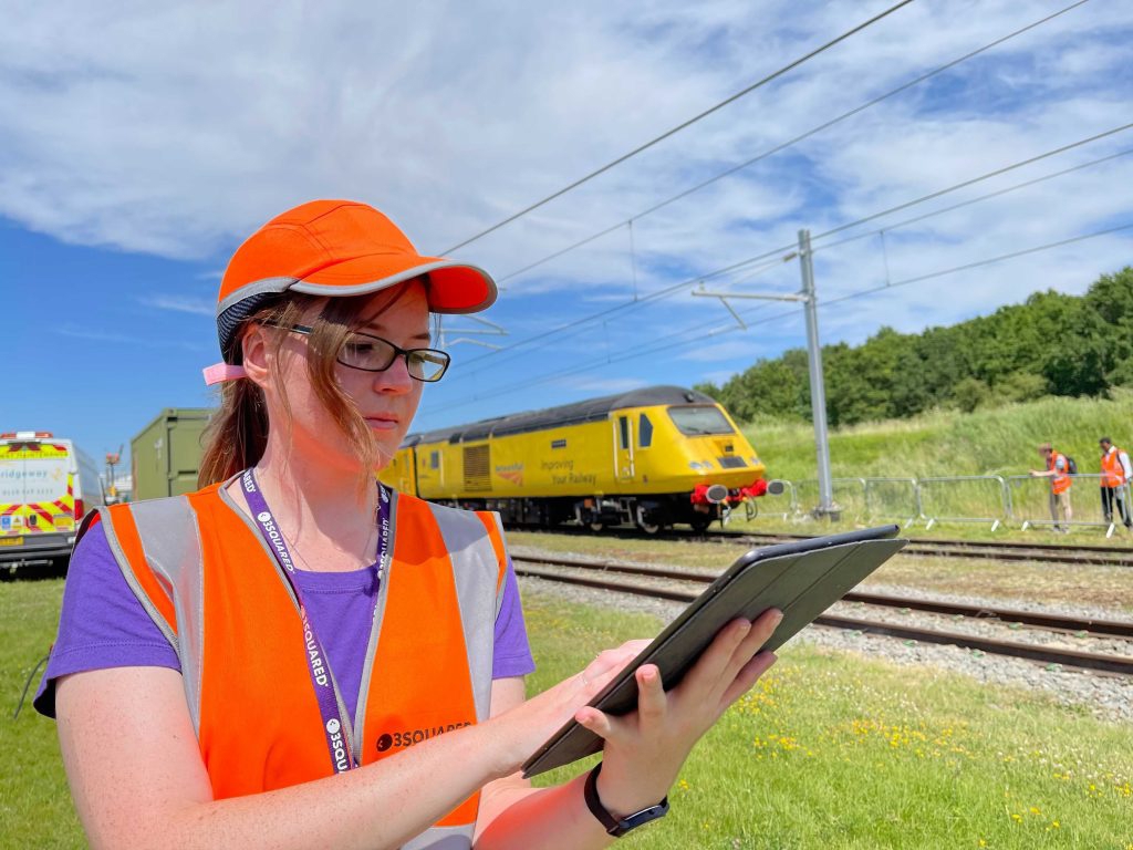 A woman wearing a high visibility vest and hat looking at a clipboard with a train going past in the background. 