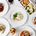 An overhead view of a table laid out with 7 plates of food and a glass of red wine.