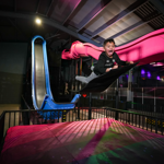 A child flies through the air towards a thick crash mat, having just come down a long slide, with a huge smile on his face.