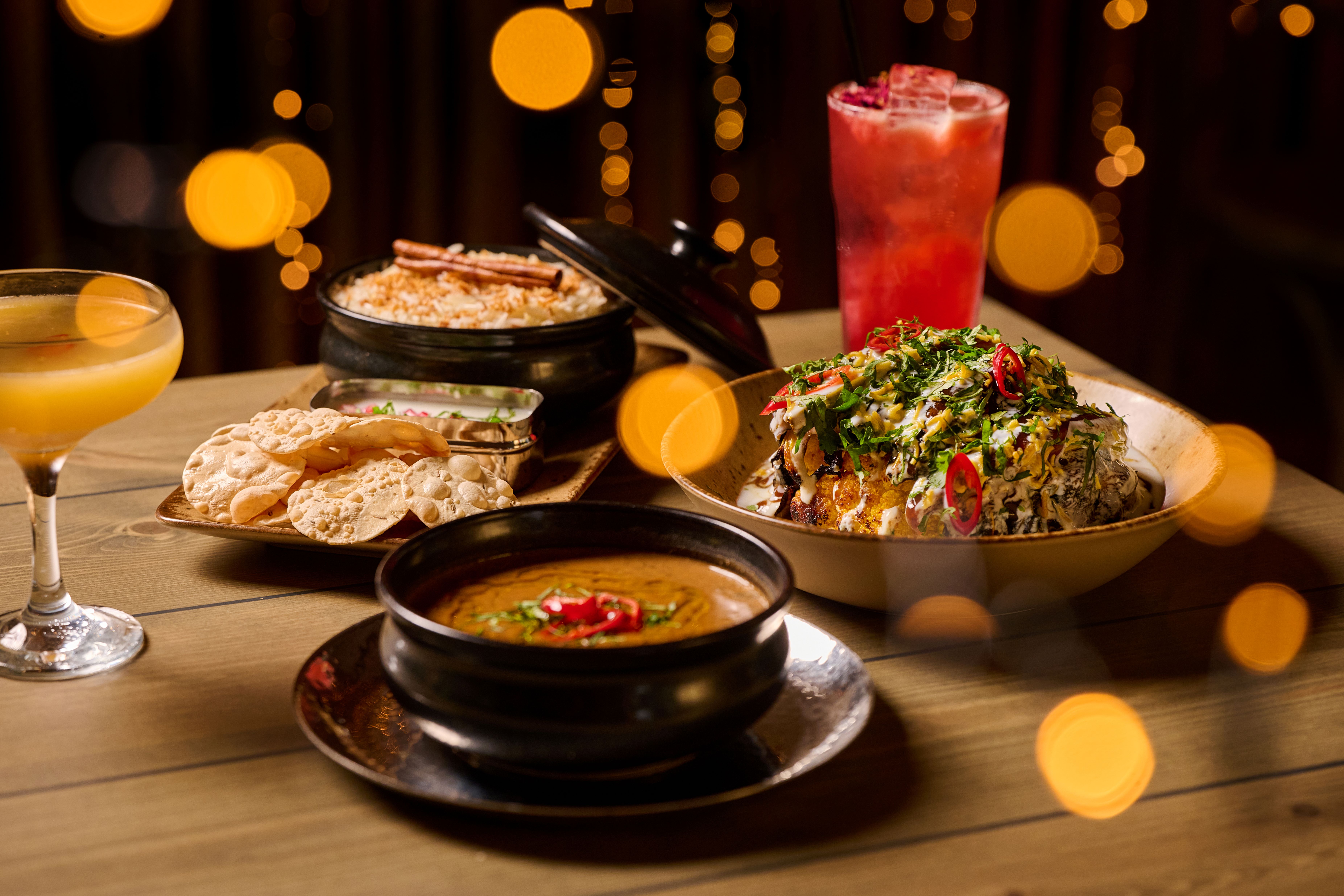 A close-up of an assortment of dishes on a wooden table, including a bowl of curry, rice, flatbreads, a spiced vegetable dish and two colourful cocktails, surrounded by warm blurred lights in the background.