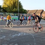 A group of people are playing Bike Polo on a tarmac area. They are riding bikes and holding polo mallets. One player is about to heat the ball. All the players are wearing helmets and protective pads.