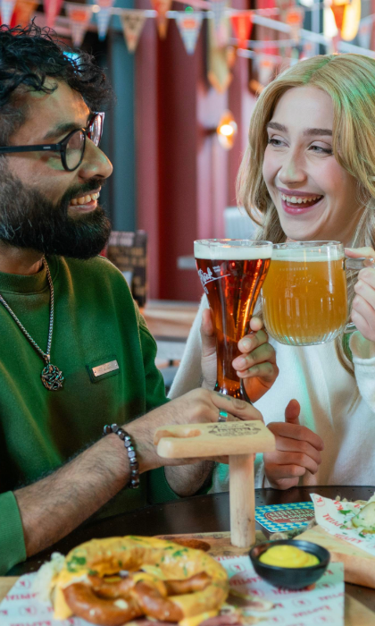 Two people are seated at a table in a beer hall, clinking glasses of beer in a toast. One glass is a tall, dark beer and the other is a large mug of pale beer. On the table are wooden boards with soft pretzels, mustard dip, and branded paper. The background features colourful bunting and warm lighting, creating a festive atmosphere.