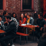 A group of people are sat at tables outside, at night, chatting and having a drink.