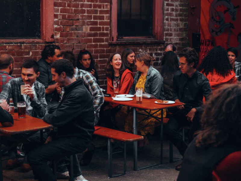 A group of people are sat at tables outside, at night, chatting and having a drink.