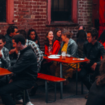 A group of people are sat at tables outside, at night, chatting and having a drink. 