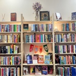 Interior view of Juno Books featuring three tall wooden bookshelves filled with neatly arranged colourful books. The centre shelf includes a small display area decorated with a crochet-style banner reading ‘JUNO’, surrounded by featured books. Dried flowers and small decorative items sit on top of the shelves, creating a warm, inviting atmosphere.