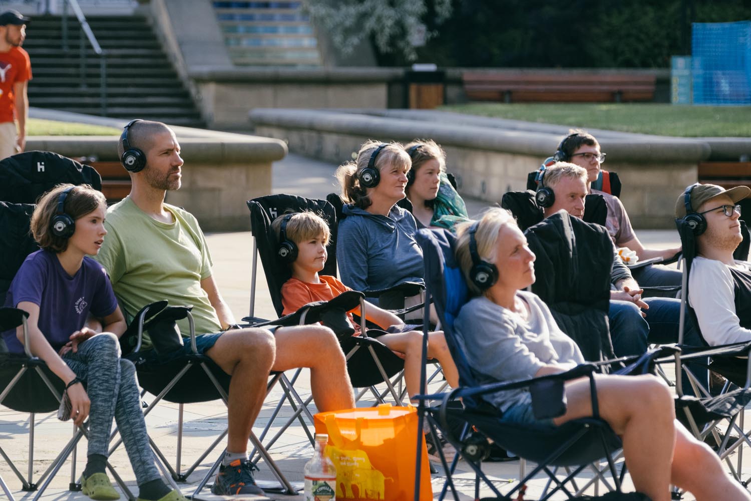 A group of people seated in folding chairs outdoors, all wearing headphones and facing the same direction as if participating in a silent event or performance. The setting appears to be a public square with stone steps, benches, and greenery in the background. A bright orange bag and a drink bottle are visible near the front row, adding colour to the scene.