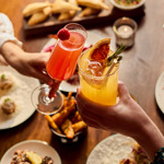 Two hands hold colourful cocktails above a table filled with delicious food.