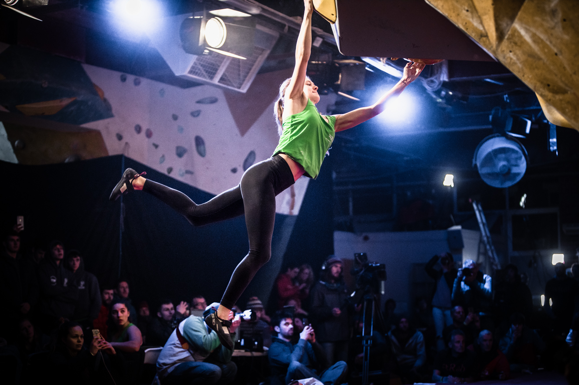 Indoor climbing competition scene with a climber hanging from an overhanging wall, wearing a green top and black leggings. One arm is extended to grip a hold while the other arm reaches upward, and one leg is stretched out for balance. Bright spotlights illuminate the climber, and a crowd of spectators is visible in the background.