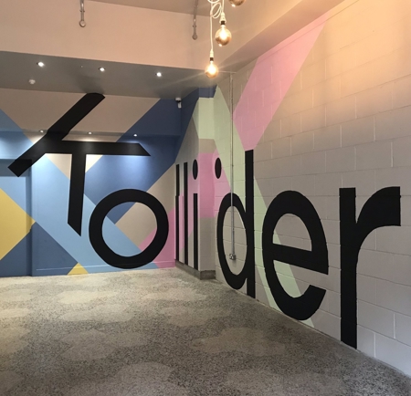 Interior view of a modern hallway with polished terrazzo flooring and colorful geometric wall art. The mural features large black letters spelling “Kollider” across a background of pastel shapes in pink, blue, yellow, and green. Several hanging light bulbs with exposed cords are suspended from the ceiling, adding a contemporary touch.