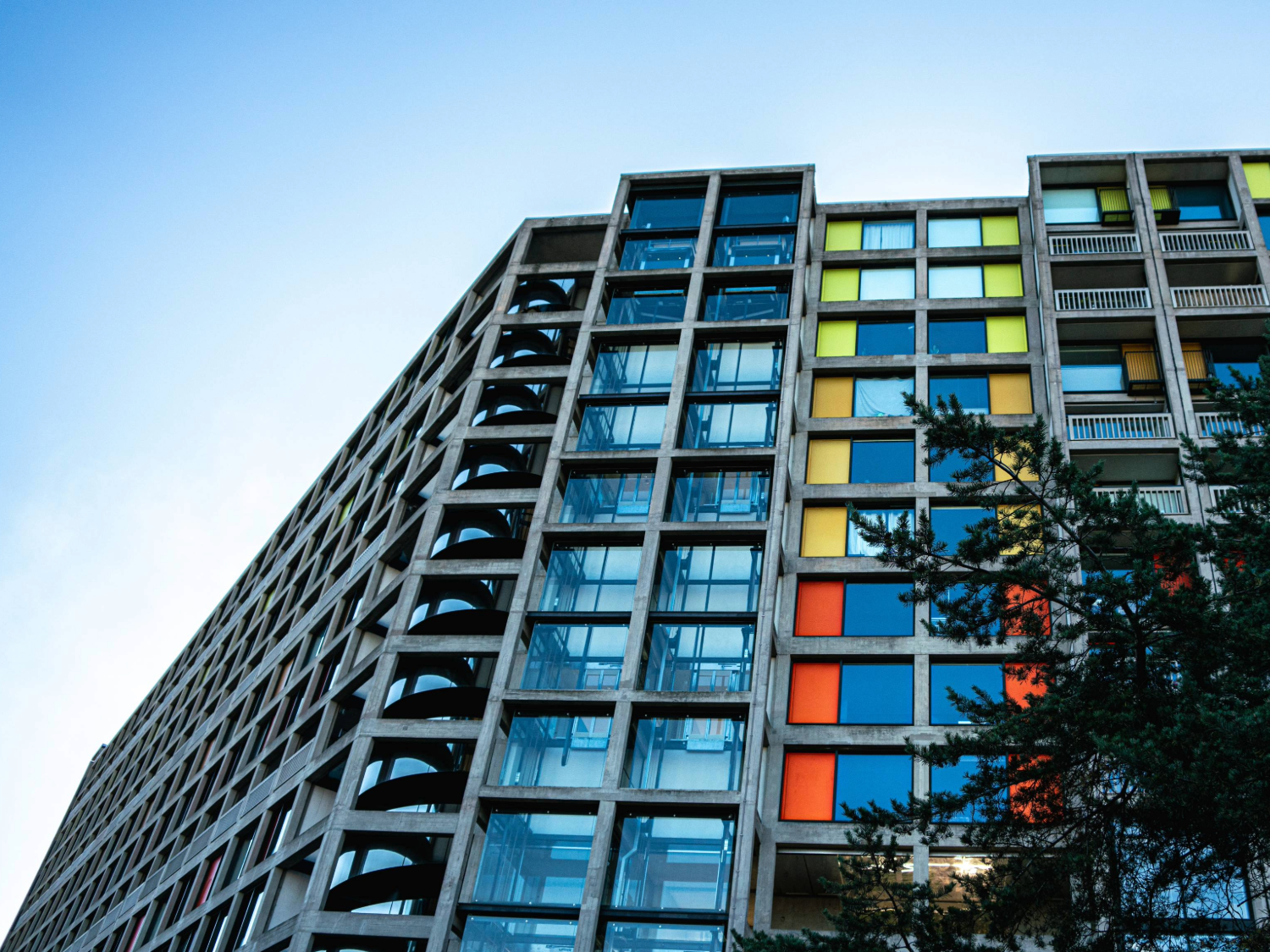 A brutalist high-rise apartment block made of concrete and glass. The windows are interspersed with bright panels in yellows and oranges.