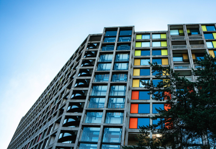 A brutalist high-rise apartment block made of concrete and glass. The windows are interspersed with bright panels in yellows and oranges.