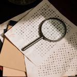 Sheets of paper covered in coded symbols lie spread out on a desk, with a magnifying glass resting on top. The papers include smudges and markings, and a few plain folders sit beneath them, creating the impression of a puzzle or secret message being studied.