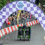 A soapbox racer decorated with artificial grass and blowing bubbles moves downhill under a large purple-and-white checkered archway. The arch displays a circular logo featuring a cartoon car and the words “Krazy Races.” Spectators line both sides of the road behind safety barriers, cheering and holding colourful flags and balloons. Trees and event signage are visible in the background.