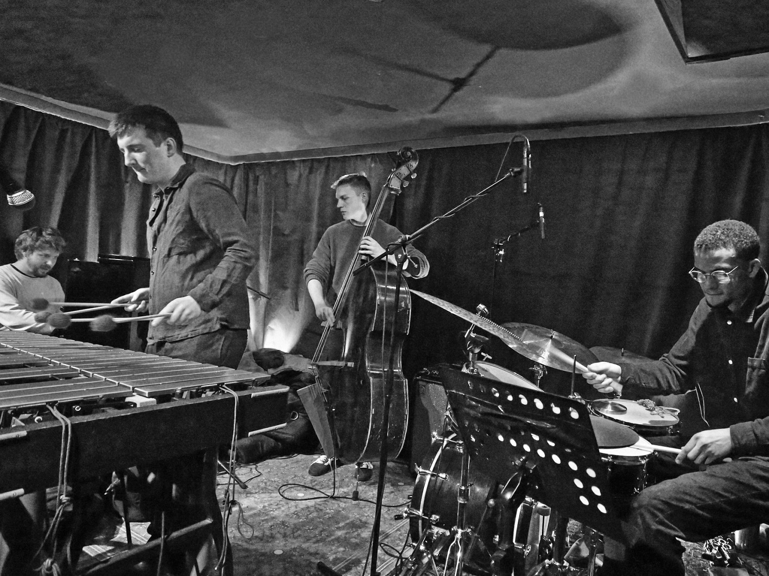 A black‑and‑white photo of four musicians performing in a small venue. A vibraphonist plays mallets on the left, with a pianist seated behind him. At the centre, a double bassist stands beside a microphone. On the right, a drummer plays a drum kit with music stands in front. Heavy curtains line the walls and equipment cables run across the floor.