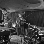A black‑and‑white photo of four musicians performing in a small venue. A vibraphonist plays mallets on the left, with a pianist seated behind him. At the centre, a double bassist stands beside a microphone. On the right, a drummer plays a drum kit with music stands in front. Heavy curtains line the walls and equipment cables run across the floor.