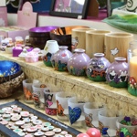 A display of colourful handmade items including painted bowls, decorated candles, illustrated vases, small trinkets, and hand‑designed cups arranged on wooden shelves.