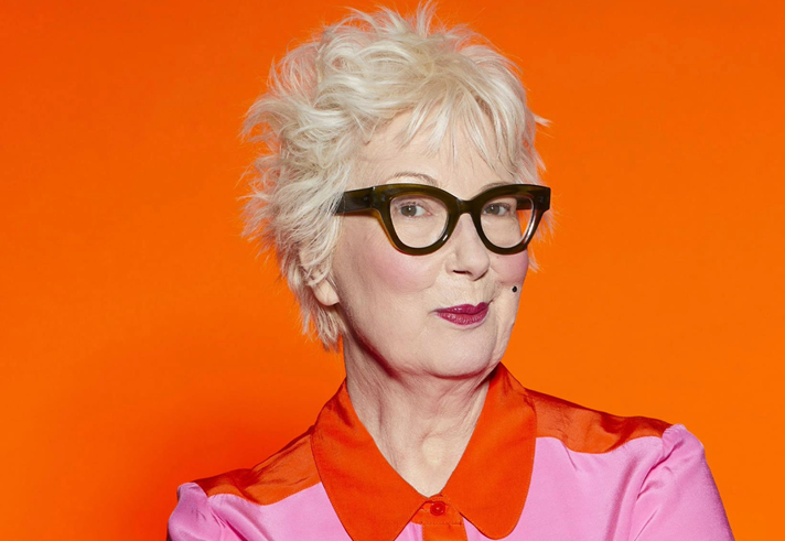 Jenny Eclair is smirking at the camera, she is wearing a pink shirt with a red collar.