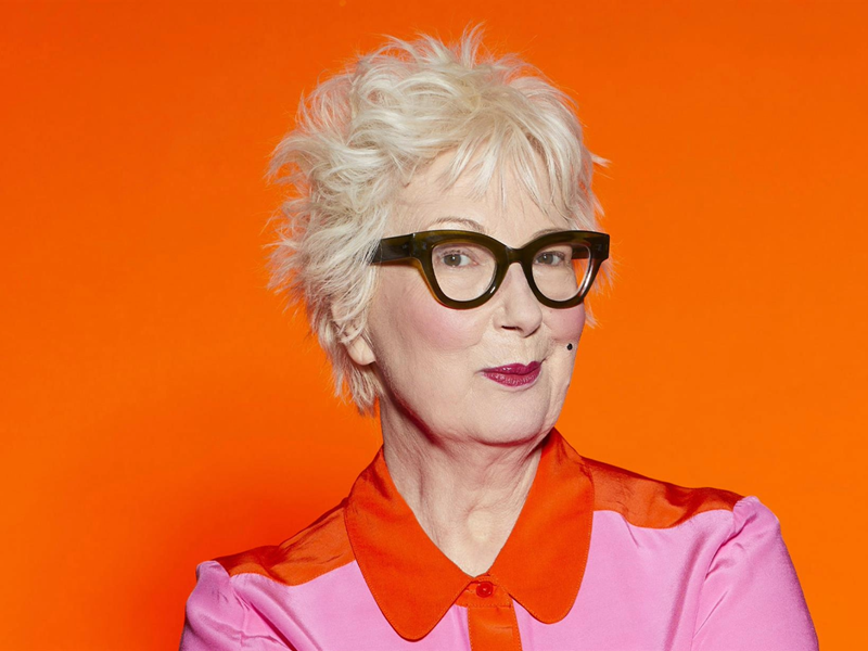 Jenny Eclair is smirking at the camera, she is wearing a pink shirt with a red collar.