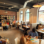 Inside The Dice Box Sheffield, there are tables and booths full of people playing games and drink and eating. The back wall is covered in shelving that is full of boxed games.
