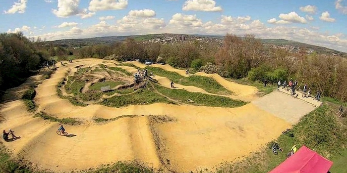 erial view of a dirt bike pump track with winding paths and raised berms surrounded by trees and greenery. Several cyclists are riding on the track, and a red canopy tent is visible in the bottom right corner. The background shows a hilly landscape under a blue sky with scattered clouds.