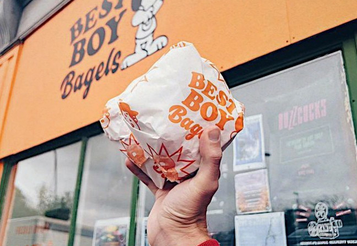 A hand holds a wrapped bagel from Best Boy Bagels, whose sign can be seen in the background.