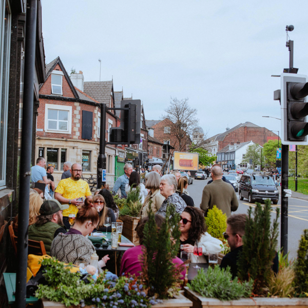 A busy street scene with people gathered at outdoor tables in front of a café or pub. The tables are decorated with planters filled with green shrubs and flowers. In the background, there are traditional brick buildings with shopfronts, a traffic light, and cars on the road. Trees and greenery are visible on the right side under a bright, overcast sky.