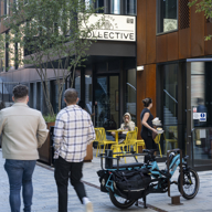 Two people walking along a modern pedestrian street with outdoor seating in front of a café called ‘Collective.’ A cargo bike is parked nearby, and a person is serving drinks to someone seated at a yellow table. The area features contemporary architecture with rust-coloured panels, large windows, and greenery.